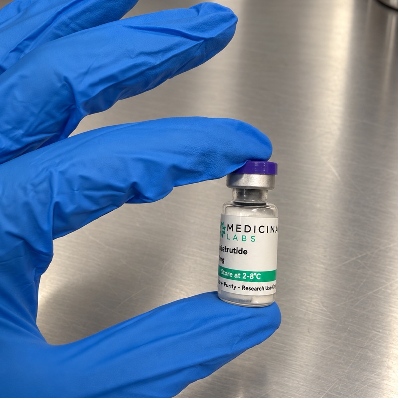 A gloved hand holding a Medicina Labs vial on a stainless steel bench